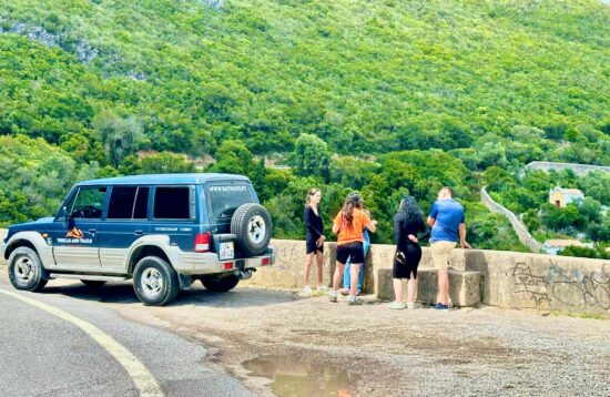 Foto de uma guia turística a explicar algo a um grupo interessado junto a um jipe 4x4, com uma vista panorâmica para o Convento da Arrábida e a serra ao fundo. A imagem transmite um momento de aprendizagem e descoberta na natureza.
